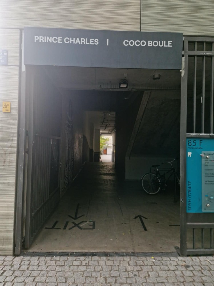 The entrance from the street: an open metal gate under a dark sign reading Prince Charles and Coco Boule. A covered passage leads straight ahead to a bright courtyard with trees.
